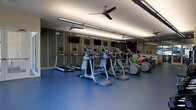 This fitness center offers a streamlined, modern workout environment designed with both function and comfort in mind. The space is outfitted with a row of cardio machines, including treadmills, ellipticals, and a stationary bike, arranged to maximize floor space and ease of movement. A large mirrored wall along one side visually expands the room while allowing users to monitor their form during exercise. The bold blue rubber flooring provides durable, slip-resistant footing suited to high-traffic athletic use. Overhead, linear LED lighting runs the length of the ceiling, delivering consistent, bright illumination throughout the space. Ceiling fans are mounted above, promoting air circulation and comfort during workouts. A textured, vertically striped accent wall adds a subtle design element, lending the space a polished aesthetic. Wall-mounted televisions are positioned for easy viewing from the cardio equipment. A free weights and stability ball area is visible near the mirrored wall, rounding out the available exercise options. Overall, the fitness center balances practicality with a clean, contemporary design.