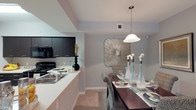This single-floor residence showcases an open-concept layout that seamlessly connects the kitchen and dining areas, creating a cohesive and functional living environment. The kitchen is appointed with dark, flat-front cabinetry that provides a striking contrast against the light-toned countertops and neutral gray walls. Black appliances, including a range and microwave, complement the kitchen's contemporary aesthetic, while a double-basin sink with a modern faucet adds practicality to the space.

The transition into the dining area is marked by a stylish pendant light suspended from the ceiling, its white dome shade casting a warm, focused glow over the dining table below. A decorative sunburst wall accent serves as a focal point, adding texture and visual interest to the dining space. The dining area comfortably accommodates a substantial wooden table paired with patterned upholstered chairs, lending an air of sophistication.

Neutral gray walls and light-toned flooring run throughout, contributing to a clean, modern ambiance. Subtle architectural details, including tray ceiling elements, add dimension and a refined finish to the overall interior.