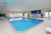 This single-level indoor pool facility is designed with both leisure and functionality in mind. The centerpiece is a generously sized rectangular lap pool with vivid blue water, lane dividers, and pool entry steps. Vaulted ceilings rise above the pool area, lending an open, airy atmosphere enhanced by recessed lighting and multiple large windows that invite natural light and exterior views.

Along one side of the pool, a row of contemporary white lounge chairs with teal accent pillows provides ample seating for relaxation. Additional lounge seating lines the opposite side, along with a small outdoor-style table and umbrella setup near the far end. A tropical beach mural decorates one wall, adding a resort-inspired touch to the environment.

A compact hot tub is located at one end of the facility, offering a dedicated space for hydrotherapy. The surrounding deck features a smooth concrete finish with tile accents bordering the pool edge. On one side of the facility, a women's restroom and additional service doors provide convenient amenities. The overall color palette of soft blue-gray walls paired with teal accents creates a clean, cohesive aesthetic throughout the space.