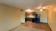 This single-floor residence presents an open-concept layout that seamlessly connects the living area to the kitchen, creating a cohesive and functional living space. The kitchen is well-appointed with dark wood cabinetry providing ample storage, a stainless sink, and a full suite of black appliances including a refrigerator, dishwasher, and range. Under-cabinet and ceiling-mounted lighting illuminate the kitchen workspace effectively. The living area features warm hardwood-style flooring that transitions into plush carpeting, adding comfort underfoot. A ceiling fan with a light fixture is centrally positioned, providing both air circulation and ambient lighting throughout the open space. The textured ceiling adds a classic architectural detail, while the neutral wall tones create a versatile backdrop suitable for a variety of décor styles. The layout makes efficient use of the available space, with a clear division between the kitchen and living zones while maintaining an airy, open feel. Overall, this residence offers a practical and inviting atmosphere, well-suited for everyday living.