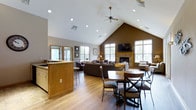 This single-floor space makes an immediate impression with its dramatic vaulted ceiling, which rises to a sharp peak and is anchored by a centrally mounted ceiling fan. The open-concept layout seamlessly connects multiple functional zones, creating a welcoming and versatile environment. Recessed lighting runs throughout the ceiling, complementing the generous natural light that streams in through numerous large windows along the walls.

The living area is anchored by a tile-surround fireplace topped with a wall-mounted television, flanked by windows that frame views of the surrounding greenery. Comfortable seating is arranged to encourage conversation and relaxation in this warm, well-lit zone. An accent wall in a rich brown tone adds depth and contrast against the crisp white of the vaulted ceiling.

A dining area with a round table and cushioned chairs occupies the foreground, transitioning naturally to a kitchen bar or service counter finished in warm wood cabinetry. Light hardwood flooring flows throughout, unifying the space and adding to its bright, airy feel. The overall ambiance is casual yet polished, making this an ideal space for gathering, socializing, or simply unwinding in a comfortable, thoughtfully designed environment.