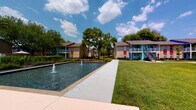 Located at 8787 Hammerly Blvd in Houston, Texas, this eye-catching residential community makes a bold architectural statement with its vividly colored building exteriors — featuring striking accents of red, blue, yellow, and teal set against neutral masonry facades. The community is thoughtfully designed around a central outdoor amenity area that serves as its social heart.

At the center of the grounds lies a generously sized swimming pool with crystal-clear turquoise water, reaching a depth of 8 feet. The pool deck is equipped with modern lounge chairs for sunbathing, and a vibrant cobalt-blue shade structure with a sleek white frame provides a stylish, sheltered retreat for residents. Palm trees and manicured greenery frame the pool area, enhancing the resort-like atmosphere.

A decorative reflecting pond with active water fountains adds an elegant focal point to the landscaped courtyard, complemented by well-maintained lawns, walkways, and mature trees that contribute to a lush, welcoming environment. Wrought-iron fencing neatly defines the pool area while maintaining open sightlines across the grounds.

The multi-building layout offers a sense of community and openness, with exterior staircases and private balconies visible throughout. The overall ambiance is lively, colorful, and inviting, reflecting a well-maintained property designed for comfortable outdoor living in the Houston climate.