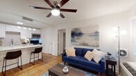 This single-floor unit offers a well-organized layout connecting the kitchen, living area, bedroom, and bathroom in a cohesive flow. The kitchen is equipped with white shaker cabinetry, granite countertops, a subway tile backsplash, and stainless steel appliances including a refrigerator, range, and over-the-range microwave. A peninsula with seating provides a casual dining option and visually connects the kitchen to the living area. The living space is comfortably sized, featuring a ceiling fan with a light fixture, neutral white walls, and warm hardwood-style flooring that carries throughout the unit. The bedroom accommodates a full-sized bed and includes a ceiling fan, a window with blinds for natural light, and direct access to the living area. One wall in the bedroom features an exposed painted concrete block finish, adding a subtle industrial character. The bathroom is clean and functional, with a tub-shower combination, a granite-topped vanity, and continued hardwood-style flooring. Exposed block tile walls in the bathroom echo the bedroom's texture. Overall, the unit maintains a bright, neutral palette with updated finishes throughout.