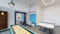 This single-floor amenity space is thoughtfully designed for social interaction and recreation, combining a lively game room with an adjacent dining area in a cohesive, modern aesthetic. The game room takes center stage with a shuffleboard table and a ping-pong table, offering guests and residents an engaging space for friendly competition. A built-in bench seat finished in vivid blue shiplap adds a nautical-inspired pop of color, complemented by a small bistro-style table and chairs for casual seating between games. Pendant lighting with glass fixtures hangs from the ceiling, casting warm, focused light throughout the space. The color palette of crisp white walls and bold blue trim creates a clean, energetic atmosphere. Through a dark-framed doorway, the adjoining dining area is visible, featuring multiple tables and chairs arranged for group gatherings, along with what appears to be a kitchenette or service counter in the background. Recessed linear lighting along the ceiling adds a contemporary touch, enhancing the brightness of the overall space. The tile flooring throughout is both practical and polished, tying the two areas together seamlessly.