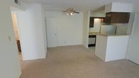 This single-floor space offers a clean, functional layout designed for comfortable everyday living. The main living area is finished with soft, neutral-toned wall-to-wall carpeting that creates a warm and inviting atmosphere, while white walls throughout lend a bright, airy feel to the interior.

A ceiling fan with a light fixture is centrally positioned overhead, providing both air circulation and ambient lighting to the living space. The open layout flows naturally into the kitchen area, which is divided by a raised breakfast bar countertop — a practical feature that defines the two spaces while maintaining an open, connected feel.

The kitchen is equipped with dark wood cabinetry, a stainless sink, a dishwasher, and a microwave, offering a well-appointed cooking space with efficient storage. Under-cabinet lighting adds a functional touch to the kitchen workspace.

A doorway visible from the living area leads to additional rooms, suggesting a thoughtful floor plan that balances open communal space with more private areas. The textured ceiling adds a subtle architectural detail throughout. Overall, the space presents a straightforward, well-maintained interior with a neutral palette that offers a versatile canvas for a variety of interior styles.