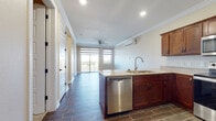 This single-floor residence offers a thoughtfully designed open-concept layout that seamlessly connects the kitchen and living areas. The kitchen is a standout feature, showcasing rich mahogany-toned cabinetry, granite-style countertops, and a full suite of stainless steel appliances including a range, over-the-range microwave, dishwasher, and refrigerator. A peninsula with an undermount sink provides additional prep space and serves as a natural divider between the kitchen and the living area. Wood-look tile flooring runs throughout the main living spaces, adding warmth and durability, while crown molding along the ceilings lends an elevated, polished finish. The living area is bright and airy, benefiting from a sliding glass door that opens to a private balcony, a ceiling fan, and a wall-mounted mini-split HVAC unit for year-round comfort. The bedrooms are carpeted and bathed in natural light through dual-pane windows with modern roller shades, offering a calm and comfortable retreat. The bathroom features a soaking tub with a speckled stone-surround enclosure and classic white fixtures. A hallway with multiple doors provides practical access to the various rooms, completing this well-appointed and functional living space.
