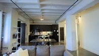 This single-story space showcases an industrial loft aesthetic through its defining architectural features. The ceiling is a standout element, displaying prominent white-painted exposed beams and structural supports that add character and a sense of vertical volume to the interior. Visible conduit and track lighting run along the ceiling, reinforcing the industrial design sensibility.

The open floor plan connects the living, dining, and kitchen areas without visual interruption. The kitchen is outfitted with dark cabinetry contrasting against stainless steel appliances, including a range, microwave, and refrigerator. A functional workspace runs along the back wall, providing ample counter and storage space.

A wooden dining table with seating sits centrally between the kitchen and living area, serving as a natural transition point between the two spaces. The living area features a light gray upholstered sofa, offering a soft contrast to the otherwise bold tones of the room.

Polished concrete floors run throughout, complementing the industrial character of the space. Structural columns and neutral white walls complete the aesthetic, creating a cohesive, contemporary environment well-suited for both living and entertaining.