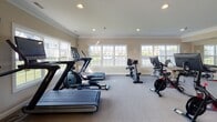 Located at 4623 Lakeside Dr in Maumee, Ohio, this fitness center offers a well-equipped, single-floor exercise facility designed to support a comprehensive workout routine. The space is finished with neutral warm-toned walls, crown molding, and carpeted flooring, giving it a clean and polished appearance.

Natural light streams in through multiple large double-hung windows positioned along the perimeter walls, providing pleasant outdoor views of the surrounding neighborhood and creating an open, inviting atmosphere. Recessed ceiling lighting supplements natural light throughout the space.

The cardio area is well-stocked with treadmills, elliptical trainers, stationary bikes, and a stair climber, catering to a wide range of cardiovascular preferences. A wall-mounted flat-screen television is conveniently positioned for entertainment during workouts. The strength training area features a multi-station cable machine, a workout bench, stability balls, and a full rack of dumbbells spanning various weight increments. A large wall mirror spans one side of the room, enhancing the sense of space and allowing users to monitor their form.

French doors with glass panes provide access to the exterior, further connecting the interior to the outdoors. The overall layout is thoughtfully organized to maximize usability, making this fitness center a functional and welcoming amenity.