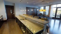 This single-floor space showcases a contemporary open-concept design that seamlessly integrates a kitchen, bar, and lounge area into one cohesive environment. The centerpiece is a generously sized kitchen island finished in light-veined granite, offering ample workspace and an integrated sink. Dark espresso cabinetry beneath the island provides strong visual contrast against the stone surface, while recessed drawers and appliances add functional depth.

A row of stylish bar seating lines one side of the island, upholstered in bold, patterned fabric that introduces warmth to the otherwise modern palette. Overhead, a series of globe pendant lights runs the length of the island, providing focused task lighting while contributing to the room's refined aesthetic.

Beyond the kitchen area, the space opens into a lounge and recreation zone featuring a billiards table and additional seating arranged around a decorative pendant chandelier with a golden shade. Dark wood paneling anchors the back wall, adding an upscale, club-like quality to the space. Natural light filters in through windows on the far wall, brightening the neutral gray tones throughout. The overall atmosphere is polished, social, and well-appointed.
