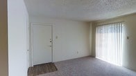This single-floor residential space offers a straightforward and functional layout designed for comfortable everyday living. The main living area is finished with wall-to-wall carpet, providing a soft and warm underfoot feel, while the entryway transitions to hardwood-style flooring, creating a practical and easy-to-clean surface near the front door. The solid white entry door features a deadbolt lock, offering a secure point of entry. Natural light fills the living space through a large window dressed with vertical blinds, allowing occupants to control light and privacy as needed. The walls are painted in a clean, neutral white tone, lending the space a bright and open feel that serves as a versatile backdrop for a variety of interior styles. The textured popcorn ceiling adds a classic architectural detail consistent with traditional residential construction. The overall ambiance is clean, simple, and move-in ready, making it a practical choice for those seeking a straightforward living environment. The open feel of the main room provides ample flexibility for furniture arrangement and personalization.