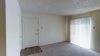 This single-floor residential space offers a straightforward and functional layout designed for comfortable everyday living. The main living area is finished with wall-to-wall carpet, providing a soft and warm underfoot feel, while the entryway transitions to hardwood-style flooring, creating a practical and easy-to-clean surface near the front door. The solid white entry door features a deadbolt lock, offering a secure point of entry. Natural light fills the living space through a large window dressed with vertical blinds, allowing occupants to control light and privacy as needed. The walls are painted in a clean, neutral white tone, lending the space a bright and open feel that serves as a versatile backdrop for a variety of interior styles. The textured popcorn ceiling adds a classic architectural detail consistent with traditional residential construction. The overall ambiance is clean, simple, and move-in ready, making it a practical choice for those seeking a straightforward living environment. The open feel of the main room provides ample flexibility for furniture arrangement and personalization.