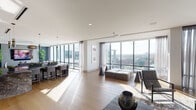 This elegant high-rise residence offers a masterfully designed open-concept layout bathed in abundant natural light. Expansive floor-to-ceiling glass windows span the length of the main living area, framing breathtaking panoramic views of a shimmering lake and a vibrant city skyline beyond — a truly captivating backdrop at every angle.

The living and entertaining areas flow seamlessly into one another, anchored by wide-plank light hardwood flooring that lends warmth throughout. A statement kitchen island clad in rich, veined stone serves as a natural focal point, complemented by sleek pendant lighting and bar seating. The kitchen features sophisticated cabinetry with integrated appliances, maintaining a clean and refined aesthetic.

The living area is generously proportioned, accommodating multiple seating arrangements beneath a striking sculptural pendant light fixture. Recessed lighting and smooth white ceilings add to the bright, airy atmosphere. A dining area with upholstered seating and a secondary lounge zone with a deep teal accent wall and lush greenery create distinct yet connected spaces.

Throughout, the interior design palette is contemporary and understated, combining neutral tones with carefully curated furnishings and artwork. The space exudes a polished, upscale ambiance that is both functional and visually impressive, with the ever-present lakeside cityscape serving as a dramatic living canvas.