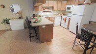 This single-floor residence offers a well-designed layout centered around a practical and inviting kitchen. The kitchen is outfitted with light wood cabinetry that provides ample storage, complemented by neutral-toned countertops that extend into a peninsula-style breakfast bar with seating for two. This casual dining feature serves as a natural divider between the kitchen and the adjacent carpeted living area, creating a sense of openness while maintaining distinct zones. The kitchen floor transitions to warm light hardwood, adding a clean and classic contrast to the surrounding carpeting. A full suite of white appliances — including a refrigerator, stove, dishwasher, and over-the-range microwave — is neatly integrated into the layout, maximizing both function and efficiency. The sink is positioned beneath a window, allowing natural light to brighten the workspace. The overall ambiance is warm and approachable, with a neutral color palette throughout that lends itself well to a variety of décor styles. The single-floor plan ensures easy accessibility and a cohesive flow throughout the entire space.