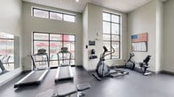 Located at 1629 S Razorback Rd in Fayetteville, Arkansas, this dedicated fitness center offers a comprehensive workout environment designed to accommodate a wide range of exercise needs. The space features soaring ceilings with a drop-tile finish and large floor-to-ceiling windows framed in black steel, flooding the interior with natural light and offering views of the surrounding area. The durable dark rubber flooring provides a practical, gym-appropriate surface throughout.

Cardio equipment includes multiple treadmills and an elliptical trainer, all positioned near the expansive windows to take advantage of the exterior views. A stationary bike adds to the variety of aerobic options available. On the strength training side, a multi-station cable and weight machine provides versatile resistance workout capabilities, complemented by a full rack of dumbbells ranging across various weights. Stability balls, medicine balls, and exercise mats round out the functional fitness offerings.

A large wall-mounted mirror spans one side of the room, enhancing the sense of openness and allowing users to monitor their form. A wall-mounted flat-screen television provides entertainment during workouts. The space is accessed through glass double doors and is finished with neutral gray walls, creating a clean, modern aesthetic. A ceiling fan provides additional air circulation, contributing to a comfortable workout environment year-round.