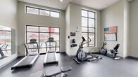 Located at 1629 S Razorback Rd in Fayetteville, Arkansas, this dedicated fitness center offers a comprehensive workout environment designed to accommodate a wide range of exercise needs. The space features soaring ceilings with a drop-tile finish and large floor-to-ceiling windows framed in black steel, flooding the interior with natural light and offering views of the surrounding area. The durable dark rubber flooring provides a practical, gym-appropriate surface throughout.

Cardio equipment includes multiple treadmills and an elliptical trainer, all positioned near the expansive windows to take advantage of the exterior views. A stationary bike adds to the variety of aerobic options available. On the strength training side, a multi-station cable and weight machine provides versatile resistance workout capabilities, complemented by a full rack of dumbbells ranging across various weights. Stability balls, medicine balls, and exercise mats round out the functional fitness offerings.

A large wall-mounted mirror spans one side of the room, enhancing the sense of openness and allowing users to monitor their form. A wall-mounted flat-screen television provides entertainment during workouts. The space is accessed through glass double doors and is finished with neutral gray walls, creating a clean, modern aesthetic. A ceiling fan provides additional air circulation, contributing to a comfortable workout environment year-round.