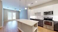Located at 224 Newell Ave in San Antonio, TX, this one-bedroom, one-bathroom unit offers a well-designed single-floor layout that maximizes its compact footprint. The open-concept kitchen and living area form the heart of the space, anchored by a large center island with a white quartz countertop and built-in shelving below, illuminated by stylish pendant lighting. The kitchen is equipped with stainless steel appliances — including a range, microwave, dishwasher, and a side-by-side refrigerator with ice and water dispenser — complemented by white shaker upper cabinets, gray lower cabinets, and a classic subway tile backsplash. Light hardwood-style flooring runs throughout the main living areas, adding warmth and continuity. The living room benefits from natural light through French doors. The bedroom features plush carpet flooring, a ceiling fan, and access to a dedicated closet with built-in shelving and a hanging rod for organized storage. The bathroom is finished with wood-look flooring, a white vanity with a gray countertop, a large mirror, and a glass-enclosed walk-in shower with white tile surround. Neutral gray tones throughout lend a clean, modern aesthetic to the entire space.