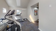 This dedicated fitness facility is designed to support a comprehensive workout experience within a thoughtfully laid-out, single-floor space. The main workout area features soaring vaulted ceilings that lend an open, airy feel, enhanced by a row of skylights that flood the room with natural light. Multiple clerestory windows along the upper walls bring in additional daylight while maintaining privacy, and a wall-mounted mini-split system ensures year-round climate comfort.

The cardio section is outfitted with a selection of Matrix brand equipment, including treadmills, an elliptical trainer, and a recumbent bike, all arranged to allow ample space between machines. The flooring throughout is durable, low-pile carpet tile in a neutral gray tone, providing cushioning underfoot while remaining easy to maintain.

A dedicated strength-training alcove houses a Hoist Mi6 functional training system, a versatile cable machine paired with an adjustable bench, offering a wide range of resistance exercises. This recessed area is finished with the same neutral wall tones and carpet flooring as the main space.

The facility is accessible via a glass-paneled door and features clean, light-gray painted walls throughout, creating a bright and motivating environment. Framed fitness reference charts are posted for user guidance, completing a well-appointed, professional-grade gym space.