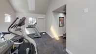 This dedicated fitness facility is designed to support a comprehensive workout experience within a thoughtfully laid-out, single-floor space. The main workout area features soaring vaulted ceilings that lend an open, airy feel, enhanced by a row of skylights that flood the room with natural light. Multiple clerestory windows along the upper walls bring in additional daylight while maintaining privacy, and a wall-mounted mini-split system ensures year-round climate comfort.

The cardio section is outfitted with a selection of Matrix brand equipment, including treadmills, an elliptical trainer, and a recumbent bike, all arranged to allow ample space between machines. The flooring throughout is durable, low-pile carpet tile in a neutral gray tone, providing cushioning underfoot while remaining easy to maintain.

A dedicated strength-training alcove houses a Hoist Mi6 functional training system, a versatile cable machine paired with an adjustable bench, offering a wide range of resistance exercises. This recessed area is finished with the same neutral wall tones and carpet flooring as the main space.

The facility is accessible via a glass-paneled door and features clean, light-gray painted walls throughout, creating a bright and motivating environment. Framed fitness reference charts are posted for user guidance, completing a well-appointed, professional-grade gym space.