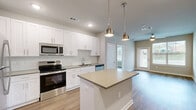 Located at 6015 Mar Dhea Lane in Charlotte, NC, this single-floor, one-bedroom, one-bathroom residence offers a thoughtfully designed layout that maximizes functionality and comfort. The open-concept living area seamlessly combines the dining room, kitchen, and living room into one cohesive space, creating an inviting atmosphere ideal for both everyday living and entertaining. The kitchen is a standout feature, showcasing white shaker-style cabinetry, a classic subway tile backsplash, stainless steel appliances including a range, microwave, dishwasher, and refrigerator, and a central island with pendant lighting above — providing both additional prep space and a casual dining option. Warm-toned wood-look flooring runs throughout the home, lending a clean, consistent aesthetic. The living area benefits from natural light through large windows and a glass exterior door, offering pleasant outdoor views. The bedroom is a comfortable, well-proportioned retreat with a ceiling fan, natural light, and direct access to the full bathroom and a spacious walk-in closet with wire shelving. A dedicated utility room houses a stacked washer and dryer, adding practical convenience. Neutral wall tones and clean finishes throughout give the space a bright, modern feel.