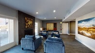 This well-appointed single-floor community space offers a welcoming and stylishly designed environment suited for relaxation and socializing. The main lounge area is anchored by a sleek, dark-tiled fireplace that serves as a striking focal point, flanked by large glass doors that allow natural light to fill the room and provide views to the outdoors. A generous arrangement of plush blue velvet armchairs surrounds marble-topped coffee tables, creating an inviting seating area ideal for casual gatherings.

The flooring throughout is a durable, wood-look plank that lends warmth and continuity to the space. Recessed lighting illuminates the room evenly, complementing the natural light. Curated wall art, including an abstract textured canvas above the fireplace and a large landscape painting, add visual interest and a refined aesthetic.

A dedicated kitchenette area features white shaker-style cabinetry, a stainless steel refrigerator, an under-mounted sink, and open shelving with decorative accents — providing practical amenities for events and everyday use. A game board display and a small café-style seating area with a bistro table and chairs round out the space, offering multiple zones for different activities.

The entrance features a set of glass double doors, creating a bright and open transition into the lounge. Overall, the space balances modern design with functional, community-focused amenities.
