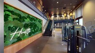 Located at 50 S Grant Ave in downtown Columbus, this two-story fitness facility delivers a well-appointed, design-forward environment across multiple dedicated zones. The main level makes an immediate impression with a large living moss wall framed in warm wood, accented by a neon "hustle" sign, setting an energetic yet refined tone. Polished concrete floors and recessed lighting carry throughout the main workout floor, which houses a comprehensive array of strength and cardio equipment, including resistance machines and free weights arranged beneath soaring ceilings with floor-to-ceiling windows that invite natural light and street-level views of the surrounding cityscape. Large-scale athletic photography adorns partition walls, reinforcing the facility's motivational aesthetic. A dedicated studio space features warm hardwood flooring, a wall-mounted display screen, full-length mirrors, and striking oversized wall murals, creating an immersive environment for group fitness or individual practice. A staircase with gold-accented railings leads to the upper level, where spin bikes and treadmills are positioned alongside open mezzanine railings overlooking the main floor below. A well-designed locker corridor features dark marble-look tile flooring, warm wood millwork, built-in vanity stations with illuminated mirrors, and built-in bench seating, providing a polished, hotel-quality finish throughout.