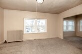 This 2-bedroom, 1-bathroom single-floor unit offers a straightforward and functional layout that includes two bedrooms, a kitchen, a bathroom, and a hallway. The primary bedroom is the largest room in the unit, featuring plush carpet flooring, crown molding, and multiple windows that invite natural light and provide views of the surrounding neighborhood's rooftops and mature trees. A classic cast-iron radiator provides heating, adding a period-appropriate architectural detail. The second bedroom mirrors a similar character, with carpet flooring, crown molding, and large windows offering comparable outdoor views. A central hallway connects the rooms, with traditional wood-panel doors adding a touch of vintage charm throughout. The kitchen features tile flooring, a large window that brightens the space, wood-toned cabinetry, a gas range, a sink, and a full-size refrigerator. The bathroom includes a full bathtub with a tiled surround, a wood-vanity with an integrated sink, a medicine cabinet mirror, and tile flooring. Overall, the unit presents a straightforward residential character with classic architectural details and consistent natural light throughout.