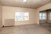 This 2-bedroom, 1-bathroom single-floor unit offers a straightforward and functional layout that includes two bedrooms, a kitchen, a bathroom, and a hallway. The primary bedroom is the largest room in the unit, featuring plush carpet flooring, crown molding, and multiple windows that invite natural light and provide views of the surrounding neighborhood's rooftops and mature trees. A classic cast-iron radiator provides heating, adding a period-appropriate architectural detail. The second bedroom mirrors a similar character, with carpet flooring, crown molding, and large windows offering comparable outdoor views. A central hallway connects the rooms, with traditional wood-panel doors adding a touch of vintage charm throughout. The kitchen features tile flooring, a large window that brightens the space, wood-toned cabinetry, a gas range, a sink, and a full-size refrigerator. The bathroom includes a full bathtub with a tiled surround, a wood-vanity with an integrated sink, a medicine cabinet mirror, and tile flooring. Overall, the unit presents a straightforward residential character with classic architectural details and consistent natural light throughout.