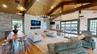 Located at 988 Old Shirley Rd in Central, SC, this single-story recreational space is a masterfully designed gathering area that blends rustic character with modern comfort. The space is defined by its dramatic vaulted ceiling clad in warm natural wood planks, supported by exposed heavy timber beams that add structural elegance and architectural depth throughout.

The open-concept floor plan is anchored by a sleek linear fireplace set into a stone accent wall, above which a large flat-screen TV is mounted. A statement chandelier with candle-style lighting hangs at the center of the main lounge area, where plush sofas and chaise lounges are arranged over area rugs atop rich hardwood floors. Adjacent to the lounge, a pool table and foosball table define a dedicated game zone, illuminated by modern linear pendant lighting.

Floor-to-ceiling steel-framed windows and large glass panel doors—including an oversized folding garage-style door—flood the interior with natural light and provide sweeping views of the outdoor pool area and patio. The seamless indoor-outdoor connection makes this space ideal for social gatherings and leisure. Every detail, from the stone masonry to the curated furnishings, reflects a refined yet relaxed aesthetic suited for community or resort-style use.