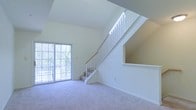 This two-floor space makes a strong first impression through its open, airy design. The main living area is defined by soaring vaulted ceilings that amplify the sense of vertical space, creating an expansive and welcoming atmosphere. Soft, neutral-toned carpet covers the floor, lending a clean and understated aesthetic throughout the lower level.

A large sliding glass door dominates one wall of the living area, allowing abundant natural light to flood the interior while offering views of the surrounding tree line, providing a pleasant connection to the outdoors.

A white-railed staircase with a warm wood handrail rises along the interior wall, leading to the second floor. An open upper landing overlooks the main living area below, reinforcing the open-concept feel between the two levels. The angled ceiling follows the roofline, contributing to the dramatic architectural character of the space.

The overall color palette is light and neutral, with white walls and trim throughout, creating a bright, clean canvas. The combination of high ceilings, natural light, and a thoughtful two-floor layout makes this a versatile and visually appealing space.