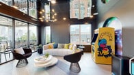 Located at 1015 N Alexander Street in Charlotte, NC, this contemporary lounge and entertainment space delivers a bold, vibrant atmosphere with a carefully curated design aesthetic. The room is defined by dramatic dark walls, reflective mirrored ceiling panels, and a striking sculptural chandelier that commands attention overhead. Large windows flood the interior with abundant natural light while offering views of the surrounding Charlotte neighborhood.

Plush, modern seating — including a tufted cream sofa accented with yellow, blue, and gray throw pillows — is arranged around a tiered circular coffee table, creating an inviting social hub. A classic Pac-Man arcade cabinet and an additional retro arcade machine, each framed by illuminated arched backdrops, add a playful, nostalgic element to the space. A sleek wet bar area features under-counter refrigeration, a sink, penny-tile backsplash, and a wall-mounted television with gold accent sconce lighting — ideal for hosting.

Floor-to-ceiling glass doors open onto a private outdoor balcony fitted with a striped pergola canopy and seating, revealing sweeping views of the Charlotte city skyline and lush tree canopy. The layered flooring, mixing light tile and linear plank patterns, ties the space together with textural contrast, making this lounge an exceptional blend of entertainment, comfort, and urban sophistication.