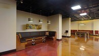 This single-floor commercial space presents a bold and contemporary interior design that balances industrial elements with refined aesthetic touches. The standout feature of the main lounge and dining area is the richly stained, high-gloss polished concrete floor in deep terracotta and amber tones, which reflects light beautifully throughout the space. Above, an exposed black ceiling adds a dramatic industrial character, with structural elements and lighting infrastructure left intentionally visible for an edgy, modern feel. A series of decorative cylindrical pendant lights hang at varying intervals, casting warm, focused illumination over the seating area. Built-in dark leather banquette seating lines one wall, paired with low wooden coffee tables featuring sculptural metal bases, creating a comfortable and inviting lounge zone. Abstract artwork adorns the warm-toned walls, adding a creative focal point to the space. Structural columns define zones without fully enclosing them, maintaining an open and airy flow. In the background, bar-height seating and red dining chairs hint at a more formal dining area, suggesting versatile use for both casual and social gatherings. Overall, the space conveys a vibrant, upscale atmosphere well-suited for a restaurant, bar, or entertainment venue.