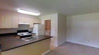 This single-floor residence offers a functional and practical layout designed for comfortable everyday living. The kitchen serves as a central hub of the space, featuring white cabinetry, a black electric range, a full-size refrigerator, and a dark-countertop peninsula with a double sink. The peninsula provides a natural division between the kitchen and the adjoining living area, creating an open-concept feel while maintaining distinct zones for cooking and relaxing. The living area is finished with neutral wall-to-wall carpeting, contributing to a warm and understated ambiance throughout. A textured ceiling adds subtle character to the interior. One of the standout conveniences of this residence is the dedicated in-unit laundry closet, visible just off the kitchen, which houses a washer and dryer — offering the practicality of on-site laundry without leaving the home. The overall color palette is light and neutral, with white walls and soft tones that create a bright, clean atmosphere. The layout is straightforward and efficient, making it easy to navigate and utilize every part of the space effectively.