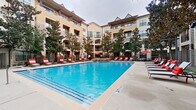 This outdoor amenity space centers around a generously sized swimming pool with crystal-clear blue water, designed to serve as a relaxing retreat within a multi-story residential community. The pool area is enclosed by a wrought-iron fence and framed by the warm stucco and stone-accented facades of the surrounding buildings, which feature arched entryways, balconies, and large windows that overlook the courtyard.

The expansive pool deck is finished in smooth concrete and lined with rows of dark-framed chaise lounge chairs dressed in bold red-and-white cushions, providing ample space for sunbathing. Vibrant red market umbrellas are positioned throughout the deck, offering shaded seating areas with outdoor furniture for lounging or socializing. Mature trees are interspersed around the perimeter, adding natural greenery and additional shade to the environment.

A standout feature of the space is a stone outdoor fireplace located along one side of the pool, creating a cozy focal point ideal for cooler evenings. The overall ambiance blends resort-style comfort with an inviting residential atmosphere, offering a well-appointed outdoor destination designed for both leisure and community gathering.