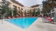 This outdoor amenity space centers around a generously sized swimming pool with crystal-clear blue water, designed to serve as a relaxing retreat within a multi-story residential community. The pool area is enclosed by a wrought-iron fence and framed by the warm stucco and stone-accented facades of the surrounding buildings, which feature arched entryways, balconies, and large windows that overlook the courtyard.

The expansive pool deck is finished in smooth concrete and lined with rows of dark-framed chaise lounge chairs dressed in bold red-and-white cushions, providing ample space for sunbathing. Vibrant red market umbrellas are positioned throughout the deck, offering shaded seating areas with outdoor furniture for lounging or socializing. Mature trees are interspersed around the perimeter, adding natural greenery and additional shade to the environment.

A standout feature of the space is a stone outdoor fireplace located along one side of the pool, creating a cozy focal point ideal for cooler evenings. The overall ambiance blends resort-style comfort with an inviting residential atmosphere, offering a well-appointed outdoor destination designed for both leisure and community gathering.