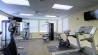 This fitness room offers a practical and comfortable environment for exercise. The space is outfitted with a diverse selection of equipment, including a FreeClimber cardio machine, a stationary bike, a multi-station cable machine, and several strength-training units, providing options for both cardiovascular and resistance-based workouts. A large wall-mounted mirror spans one side of the room, enhancing the sense of space and allowing users to monitor their form during exercise. The flooring consists of a durable, textured carpet that provides traction and cushioning underfoot, with rubber mats placed beneath heavier equipment for added protection. Natural light enters through multiple windows fitted with white horizontal blinds, creating a bright and airy atmosphere. The drop ceiling features recessed panel lighting alongside a ceiling fan, ensuring consistent illumination and airflow throughout. The walls are painted in a warm, neutral yellow tone, contributing to an inviting ambiance. Overall, the fitness room is thoughtfully arranged to accommodate a range of workout routines within a clean, well-maintained setting.