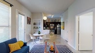 Located at 503 Avenue A in San Antonio, TX, this single-floor residence offers a thoughtfully designed open-concept layout that seamlessly connects the living, dining, and kitchen areas. The living room is a comfortable and characterful space, featuring a ceiling fan with light fixture, large windows with patterned curtains that invite natural light, and eclectic décor including framed vinyl record artwork and a bold black-and-white geometric area rug. The adjacent dining area accommodates a round white table and modern chairs beneath a globe pendant light, flowing naturally into the well-appointed kitchen. The kitchen is outfitted with dark espresso cabinetry, stainless steel appliances including a range, microwave, refrigerator, and dishwasher, along with a double sink under a window overlooking the outdoors. Polished concrete-style flooring runs throughout the main living areas, contributing to the contemporary feel of the space. The bedroom is a serene retreat with a ceiling fan, natural light from a window dressed in coordinating patterned curtains, and ample room for furnishings. The bathroom features a dark vanity with a white countertop, a large mirror, and a full tub-shower combination with white tile surround and a louvered closet door nearby.