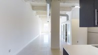 This single-floor space combines industrial and modern design elements to create a bright, versatile environment. The open layout is defined by crisp white walls and highly polished light-toned floors that reflect natural light throughout the interior. Structural columns punctuate the space at regular intervals, contributing to the industrial character while preserving an airy, unobstructed flow.

The ceiling retains its raw, exposed concrete finish, with visible ventilation ductwork running along its length — a nod to industrial-style architecture that adds visual texture against the otherwise clean palette. Recessed and track lighting supplements the abundant natural light that filters in through windows at the far end of the space, keeping the environment well-illuminated.

Along one side, glass-paneled sliding partitions create enclosed office or meeting areas while maintaining visual connectivity to the open floor. This transparency supports a collaborative atmosphere without fully sacrificing privacy. A built-in counter or workstation is partially visible, suggesting functional workspace integration. Overall, the space presents a polished, professional ambiance well-suited for office or creative studio use.