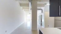 This single-floor space combines industrial and modern design elements to create a bright, versatile environment. The open layout is defined by crisp white walls and highly polished light-toned floors that reflect natural light throughout the interior. Structural columns punctuate the space at regular intervals, contributing to the industrial character while preserving an airy, unobstructed flow.

The ceiling retains its raw, exposed concrete finish, with visible ventilation ductwork running along its length — a nod to industrial-style architecture that adds visual texture against the otherwise clean palette. Recessed and track lighting supplements the abundant natural light that filters in through windows at the far end of the space, keeping the environment well-illuminated.

Along one side, glass-paneled sliding partitions create enclosed office or meeting areas while maintaining visual connectivity to the open floor. This transparency supports a collaborative atmosphere without fully sacrificing privacy. A built-in counter or workstation is partially visible, suggesting functional workspace integration. Overall, the space presents a polished, professional ambiance well-suited for office or creative studio use.