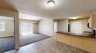 Located at 3472 4th Ave E in Dickinson, North Dakota, this two-bedroom, one-bathroom unit offers a practical and well-organized single-floor layout. The open-concept living and dining area flows seamlessly into the kitchen, creating a cohesive shared space. The kitchen is equipped with light maple cabinetry, dark granite-style countertops, a peninsula with a double sink, and black appliances including a range, microwave, and refrigerator. Tile flooring runs through the kitchen and entry areas, while plush carpet covers the living room and both bedrooms. The living room features a sliding glass door with vertical blinds that floods the space with natural light and provides access to an outdoor area, complemented by a wall-mounted air conditioning unit. Both bedrooms offer closets with paneled bi-fold doors, and the primary bedroom is a comfortable size with a window overlooking the exterior. The full bathroom includes a tub-shower combo with tile flooring and dark countertop finishes. Baseboard heating is present throughout, and a dedicated laundry area adds everyday convenience. Neutral warm-beige tones throughout give the space a clean, consistent aesthetic.