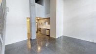 This two-story, one-bedroom, one-bathroom space showcases a distinctly industrial-modern design, blending raw architectural elements with contemporary finishes. The main living area immediately impresses with soaring double-height ceilings that create a dramatic sense of openness and volume. Exposed concrete columns anchor the space structurally while reinforcing the loft-inspired character throughout.

Polished concrete floors span the main level, offering a sleek, reflective surface that enhances the brightness of the open-plan layout. The kitchen is efficiently designed and features light wood cabinetry, a white subway tile backsplash, and a full suite of stainless steel appliances, including a refrigerator, range, microwave, and dishwasher. The clean lines of the cabinetry contrast neatly with the industrial elements present throughout the space.

Crisp white walls throughout the unit provide a neutral backdrop that complements both the raw concrete and the warm wood tones of the kitchen. The two-floor layout offers functional separation between living and private areas. Overall, the space delivers an urban loft atmosphere with modern conveniences, making it a well-balanced and visually compelling residence.