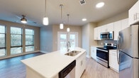 This 2-bedroom, 2-bathroom residence at The James in North Little Rock, Arkansas presents a thoughtfully designed single-floor layout that blends contemporary style with functional living. The open-concept kitchen is a standout feature, showcasing a large center island with quartz countertops, a double stainless steel sink, and stylish pendant lighting overhead. White shaker-style cabinetry extends to the ceiling, complemented by a tile backsplash and a full suite of stainless steel appliances, including a range, microwave, refrigerator, and dishwasher. French doors off the kitchen and living area invite natural light and provide outdoor access. The living area is finished with light wood-style flooring that flows throughout the unit and features a ceiling fan for added comfort. Both bedrooms are generously proportioned, with large windows offering views of the surrounding area. The primary bedroom connects to a walk-in closet and a bathroom with a tub-shower combination finished in classic white subway tile. The second bathroom features a white vanity with a large mirror and clean, neutral finishes. Warm beige tones, recessed lighting, and crisp white trim throughout create a bright, cohesive, and move-in-ready atmosphere.