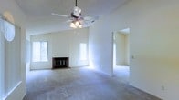 This single-floor space offers an open and airy layout defined by vaulted ceilings that lend a sense of generous vertical space to the main living area. A ceiling fan with integrated lighting is centrally mounted, providing both airflow and ambient illumination. The living room is anchored by a classic brick fireplace, adding warmth and character to the space. Neutral-toned carpet spans the floor, contributing to a soft and comfortable atmosphere throughout.

Large windows along the exterior walls invite abundant natural light, brightening the interior and enhancing the open feel of the room. The pale, neutral wall tones reflect this light effectively, keeping the space feeling fresh and welcoming.

An arched passageway connects the main living area to an adjoining room, maintaining visual continuity while subtly defining separate functional zones. The adjacent space mirrors the same carpeted flooring and light color palette, ensuring a cohesive aesthetic throughout the home. The overall design is clean and understated, offering a versatile canvas well-suited to a variety of furnishing styles and décor preferences.