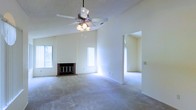 This single-floor space offers an open and airy layout defined by vaulted ceilings that lend a sense of generous vertical space to the main living area. A ceiling fan with integrated lighting is centrally mounted, providing both airflow and ambient illumination. The living room is anchored by a classic brick fireplace, adding warmth and character to the space. Neutral-toned carpet spans the floor, contributing to a soft and comfortable atmosphere throughout.

Large windows along the exterior walls invite abundant natural light, brightening the interior and enhancing the open feel of the room. The pale, neutral wall tones reflect this light effectively, keeping the space feeling fresh and welcoming.

An arched passageway connects the main living area to an adjoining room, maintaining visual continuity while subtly defining separate functional zones. The adjacent space mirrors the same carpeted flooring and light color palette, ensuring a cohesive aesthetic throughout the home. The overall design is clean and understated, offering a versatile canvas well-suited to a variety of furnishing styles and décor preferences.