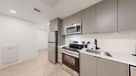 Located at 1704 North 17th Street in Tampa, FL, this modern 2-bedroom, 2-bathroom residence offers a thoughtfully designed single-floor layout with clean contemporary aesthetics throughout. Light wood-tone flooring runs seamlessly across the space, complementing a neutral palette of white and soft gray tones.

The kitchen is efficiently appointed with stainless steel appliances, including a refrigerator, electric range, over-the-range microwave, and dishwasher. Matte gray flat-front cabinetry extends to the ceiling, providing ample storage, while white countertops and a matte black faucet add a polished, modern contrast.

The living area benefits from floor-to-ceiling windows that frame views of an outdoor pool and surrounding courtyard, flooding the space with natural light. A built-in wall unit with integrated cabinetry, a dedicated workspace, and a mounted TV adds both functionality and visual cohesion.

The primary bedroom is generously sized and features direct access to one of the two full bathrooms, which are each finished with large-format tile flooring, glass-enclosed walk-in showers, and sleek matte black hardware and fixtures. Two hallways connect the rooms efficiently, contributing to a well-organized floor plan. The overall ambiance is clean, modern, and well-lit, reflecting a contemporary urban design sensibility.