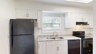 Located at 301 Village Dr in Jacksonville, NC, this two-story home offers five bedrooms and three bathrooms across two well-proportioned floors. The main level hosts the living room, kitchen, and connecting hallway, while the upper floor provides the bulk of the sleeping quarters. The living room features durable wood-look vinyl flooring, a ceiling fan, and sliding glass doors that open to a view of the outdoors, filling the space with natural light. The kitchen is equipped with white shaker-style cabinetry, light granite-look countertops, a double-basin stainless steel sink, and black appliances including a refrigerator, range, and dishwasher. A window above the sink adds brightness to the workspace. The bedrooms are carpeted and offer a clean, neutral palette, with several rooms featuring closets with built-in shelving and rods for ample storage. Natural light enters through windows in each bedroom, offering pleasant outdoor views. The bathrooms feature wood-look flooring and tub-shower combinations. Hallways connect rooms cleanly with white trim and neutral walls throughout. The home maintains a consistent, clean aesthetic with textured ceilings, white walls, and a mix of vinyl plank and carpet flooring across both levels.