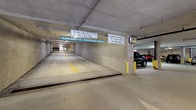This parking garage is a well-organized, multi-level facility designed to efficiently accommodate a high volume of vehicles. The structure features a concrete construction throughout, with exposed ceilings and walls that reflect the utilitarian nature of the space. Overhead and wall-mounted signage is prominently displayed, providing clear guidance to drivers, including posted clearance heights of 7 feet on the primary level and 7 feet 6 inches (6 feet 4 inches for upper level 5), along with advisories prohibiting cargo vans on certain levels. Driving lanes are clearly delineated with painted yellow directional lines and arrows on the concrete floor, ensuring smooth traffic flow through the facility. Safety-yellow bollards are strategically positioned to protect structural columns and pedestrian zones. Convex safety mirrors are installed at key junctions to improve driver visibility around corners. The garage is equipped with overhead artificial lighting that illuminates the driving lanes and parking bays. Parking spaces are arranged along the sides of the travel lanes, and the facility includes a ramp system connecting to additional parking levels. The overall layout reflects a practical, safety-conscious design intended to serve a broad range of everyday vehicle types.
