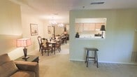 This single-floor residence showcases an open, flowing layout designed for comfortable everyday living and entertaining. The dining area serves as a focal point, anchored by an elegant chandelier that casts warm light over a generously sized dining table with seating for multiple guests. Framed artwork adorns the walls, adding a touch of refinement to the space. Soft, neutral-toned carpeting runs throughout, contributing to the overall warmth of the interior.

A defining architectural feature is the pass-through window connecting the kitchen to the adjacent living and dining areas. This opening enhances the sense of connectivity between spaces while allowing natural interaction between the kitchen and entertaining areas. The kitchen itself features light wood cabinetry, a countertop workspace, and essential appliances, all visible through the pass-through. A bar-height stool positioned at the opening provides a casual seating option.

The walls are finished in a soft sage-green tone that complements the warm beige carpeting, creating a cohesive and inviting color palette. The layout is practical and well-proportioned, offering a comfortable sense of scale without feeling overwhelming.