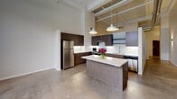 Located at 128 N Washington Ave in Minneapolis's vibrant North Loop neighborhood, this single-floor unit showcases an industrial-loft aesthetic defined by dramatically high ceilings with exposed ductwork and structural elements overhead. The open-concept layout creates a seamless flow between living and kitchen areas, anchored by polished concrete floors that reinforce the space's urban character.

The kitchen serves as a central hub of the unit, featuring a substantial center island with a clean white countertop illuminated by pendant lighting suspended from the soaring ceiling above. Dark espresso cabinetry extends to ceiling height, providing generous storage, while stainless steel appliances—including a refrigerator, electric range, and dishwasher—add a sleek, professional finish. A white subway tile backsplash completes the contemporary aesthetic.

The living area is generous and open, with exposed ventilation ducts and a baseboard heating system running along the perimeter walls, reinforcing the industrial design language. Natural light enters through windows along the outer wall.

The bathroom is thoughtfully finished with warm wood-look flooring, a dark-toned vanity, and a walk-in shower enclosed by sliding glass doors and clad in marble-style surround panels, offering an elevated, modern feel.