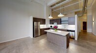 Located at 128 N Washington Ave in Minneapolis's vibrant North Loop neighborhood, this single-floor unit showcases an industrial-loft aesthetic defined by dramatically high ceilings with exposed ductwork and structural elements overhead. The open-concept layout creates a seamless flow between living and kitchen areas, anchored by polished concrete floors that reinforce the space's urban character.

The kitchen serves as a central hub of the unit, featuring a substantial center island with a clean white countertop illuminated by pendant lighting suspended from the soaring ceiling above. Dark espresso cabinetry extends to ceiling height, providing generous storage, while stainless steel appliances—including a refrigerator, electric range, and dishwasher—add a sleek, professional finish. A white subway tile backsplash completes the contemporary aesthetic.

The living area is generous and open, with exposed ventilation ducts and a baseboard heating system running along the perimeter walls, reinforcing the industrial design language. Natural light enters through windows along the outer wall.

The bathroom is thoughtfully finished with warm wood-look flooring, a dark-toned vanity, and a walk-in shower enclosed by sliding glass doors and clad in marble-style surround panels, offering an elevated, modern feel.