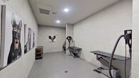 This single-level pet grooming space is designed with both functionality and cleanliness in mind. The room is finished throughout with large-format light grey wall tiles and smooth grey floor tiles, creating a hygienic, easy-to-maintain environment. Recessed ceiling vents and flush-mounted lighting provide consistent climate control and bright, even illumination throughout the space.

The grooming area is equipped with multiple stainless steel workstations, including an adjustable-height grooming table fitted with a professional-grade dryer arm and hose, as well as a dedicated wash basin station with plumbing fixtures. A floor drain is centrally positioned, facilitating efficient cleanup. A rustic industrial wood-and-metal shelving unit provides storage for grooming supplies.

The space features a private entrance through a solid grey door, ensuring secure and easy access. Decorative framed photographic prints of dogs displayed along the walls add a lively, pet-friendly character to an otherwise utilitarian setting. The layout is open and spacious, allowing comfortable movement around the equipment. Overall, the space balances a professional, sanitary design with welcoming aesthetic touches suited to a dedicated pet care facility.